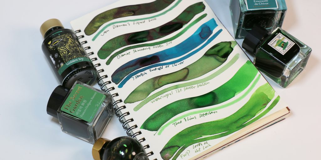 Spiral notebook with six green ink swatches featuring gold shimmer surrounded by ink the featured ink's bottles (in varying sizes). 