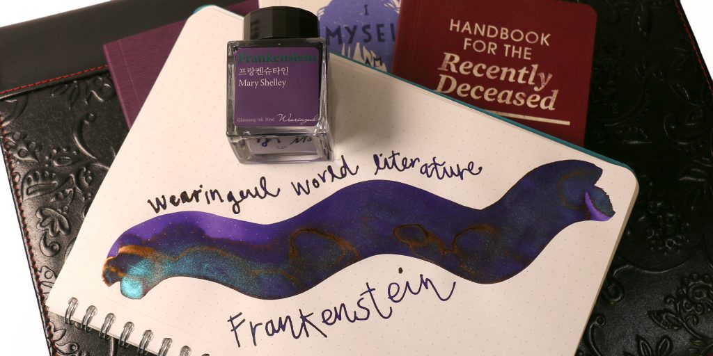Wearingeul Frankenstein ink swatch showing purple base color with pearly blue shimmer that looks like ancient moss in certain lights, and exhibiting the copper sheen you'll seen in heavy applications. Bottle of ink is beside the paint brush swatch and glass dip pen writing sample in a Wearingeul notebook. 