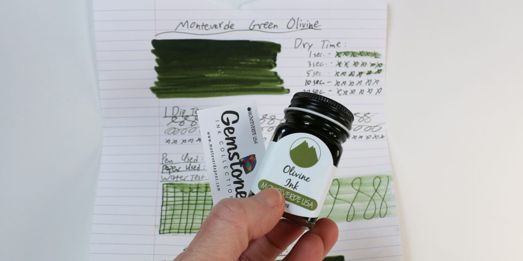 Hand holding Monteverde Olivine ink bottle with ink test sheet in background.