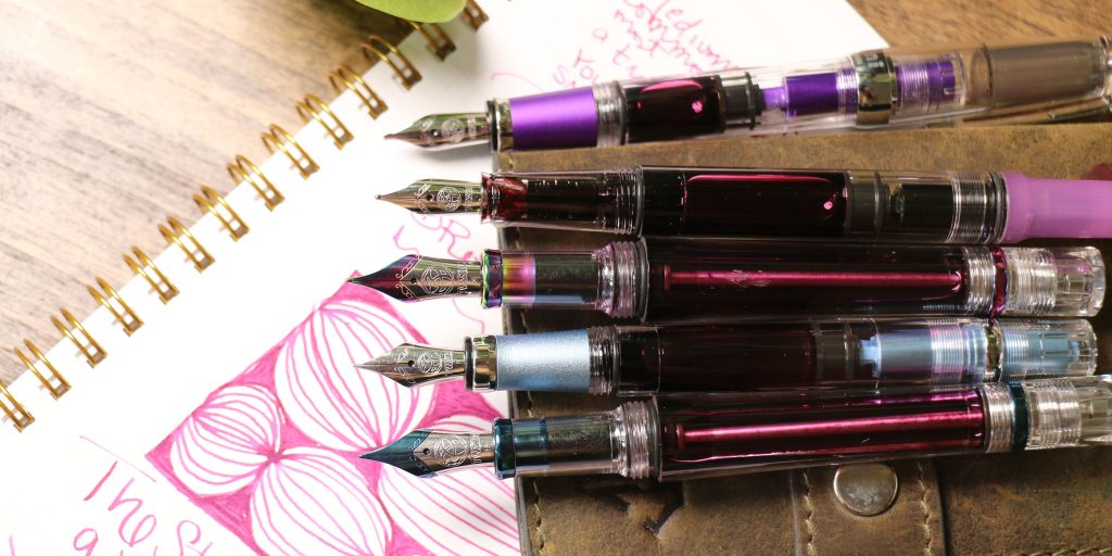 TWSBI fountain pens displayed with various nib sizes from extra fine to broad, shown on top of a notebook with ink writing samples.