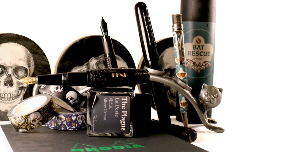 Halloween-themed writing display with fountain pens, inks, and accessories including The Plague ink, BENU Jolly Roger Pixie, Monteverde Innova, and Retro51 Bat Rescue.