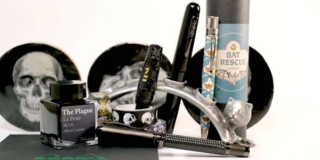 Group shot of Halloween-themed fountain pens, inks, and accessories including Wearingeul The Plague ink, BENU Jolly Roger Pixie, Monteverde Innova, Retro51 Bat Rescue, skull plates, and silver cat pen holder.