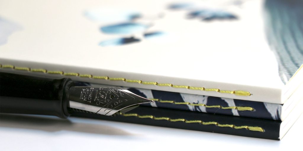 Close-up of a Benu fountain pen nib resting in front of a stack of Clairefontaine Inkebana notebooks with decorative stitching.