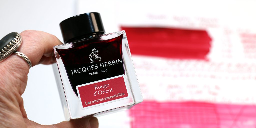 Hand holding J. Herbin Rouge d’Orient ink bottle with deep red ink and test page in background.