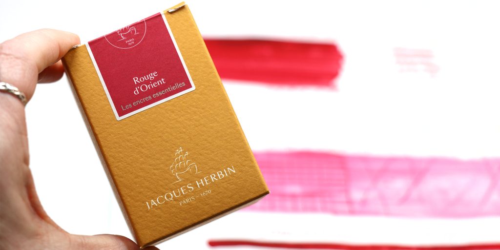 Hand holding orange J. Herbin Rouge d’Orient ink box with swatch card in background.