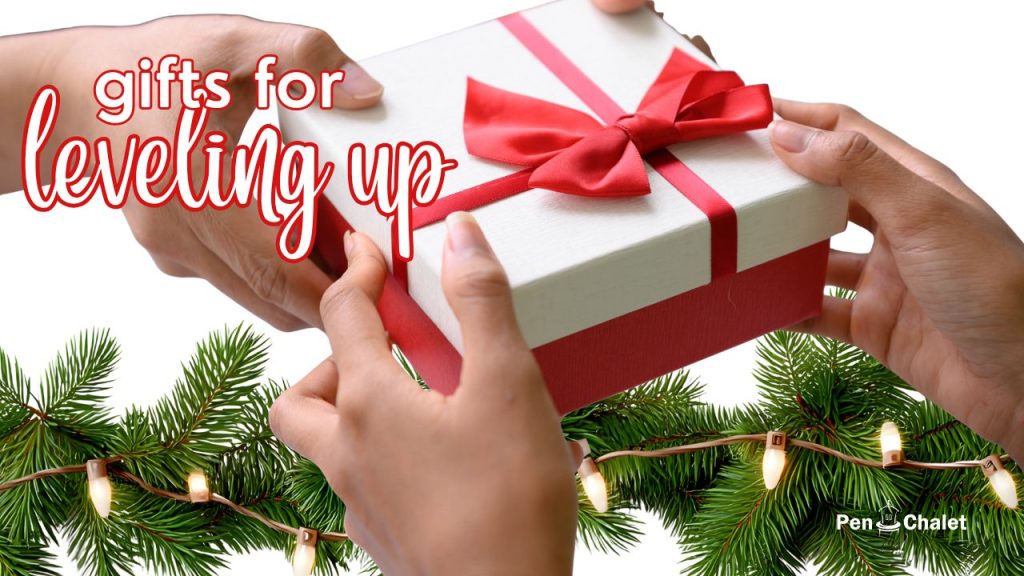 Hands exchanging a Christmas gift, white box wrapped in red ribbon in front of twinkle lights on an evergreen garland with overlay indicating a title: Gifts for Leveling Up in the 2025 Holiday Pen & Ink Gift Guide. 