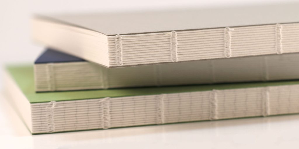 Closeup photo showing the spines/binding of the Wearingeul Reservoir Thread B6 lay flat notebooks in all three colors/paper types. 