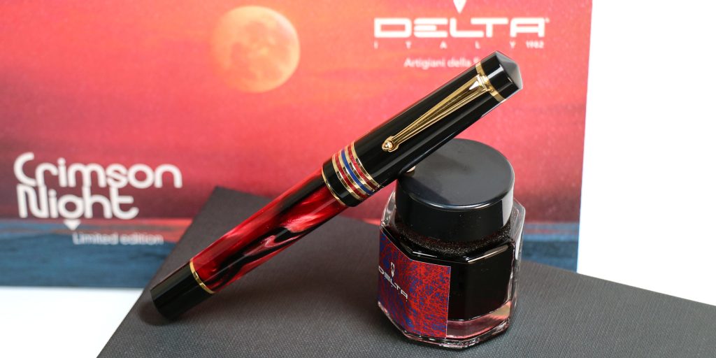 Delta Four Rings Collection Crimson Night Limited Edition fountain pen capped leaning against bottled Delta fountain pen ink in front of the custom themed sleeve for this special luxury 2026 pen release.