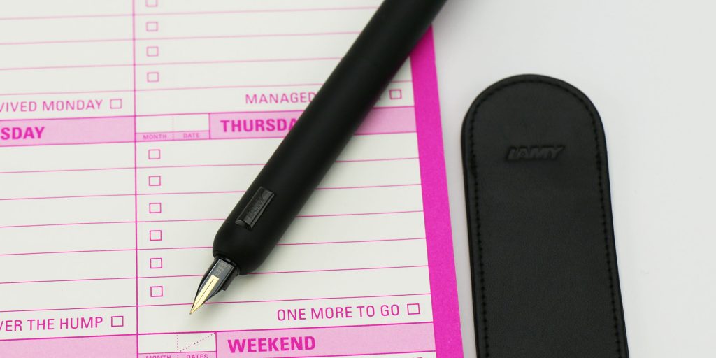 Lamy Dialog CC All Black fountain pen with retractable nib extended on top of a hot pink and white notepad for weekly planning.