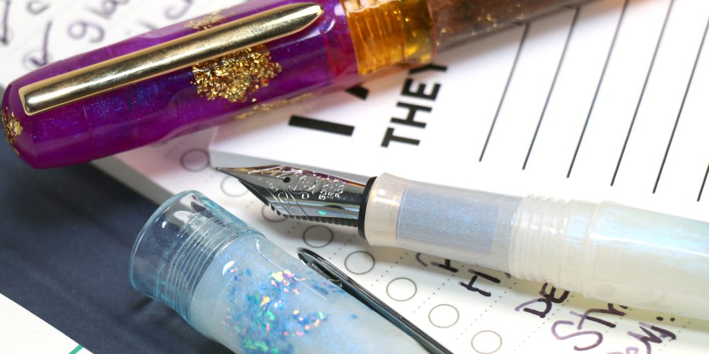 Benu Talisman Fountain pens: Ametrine and Moonstone with the Benu Moonstone Talisman fountain pen uncapped in front on pads of to do lists.