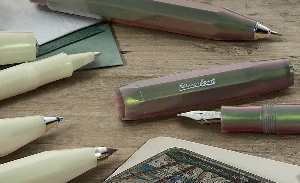 Several different Kaweco Lunar Collection pens including two colors: Shadow Green and Light Green and 3 pen modes: ballpoint, rollerball, and fountain pen open and laying in a messy grouping on a woodtop desk. 