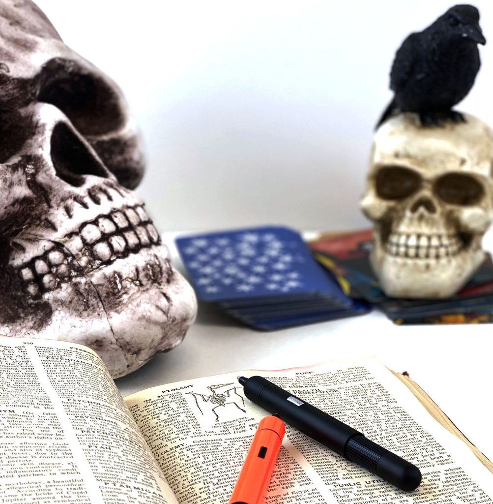 Lamy Pico black ballpoint pen in an open old dictionary with an orange Lamy Pico beside it and skulls, ravens, and tarot cards in the background.